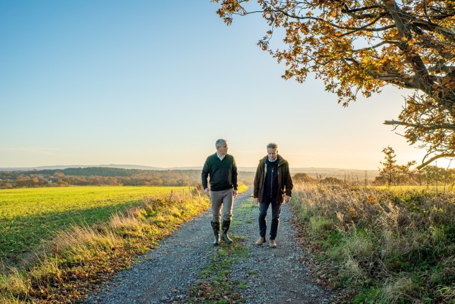 Image of two people walking in parallel along a farm track or similar, surrounded by countryside and backed by a blue sky.